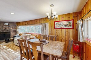 Ein Zimmer mit Holzwänden, einem Esstisch, Stühlen, einer Chandelier, einem Kamin und einem Fenster mit Vorhängen.
