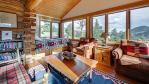A cozy cabin interior with a wooden ceiling, large windows, a fireplace, and a bookshelf. - Historic Cabin With Mountain Views, Private W/d, & Furnished Deck (Estes Park)