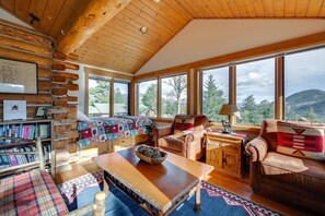 A cozy cabin interior with a wooden ceiling, large windows, a fireplace, and a bookshelf. - Historic Cabin With Mountain Views, Private W/d, & Furnished Deck (Estes Park)