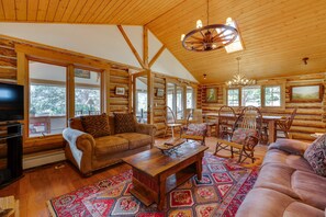 A log cabin interior with a wooden ceiling, large windows, a television, a sofa, a coffee table, and a chandelier. - Historic Cabin With Mountain Views, Private W/d, & Furnished Deck (Estes Park)