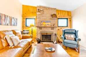 A living room with a stone fireplace, leather sofa, and a blue armchair. - Hot Tub, Pool Table, and Dog Friendly! River Waterfront Cabin With Mountain Views (Estes Park)