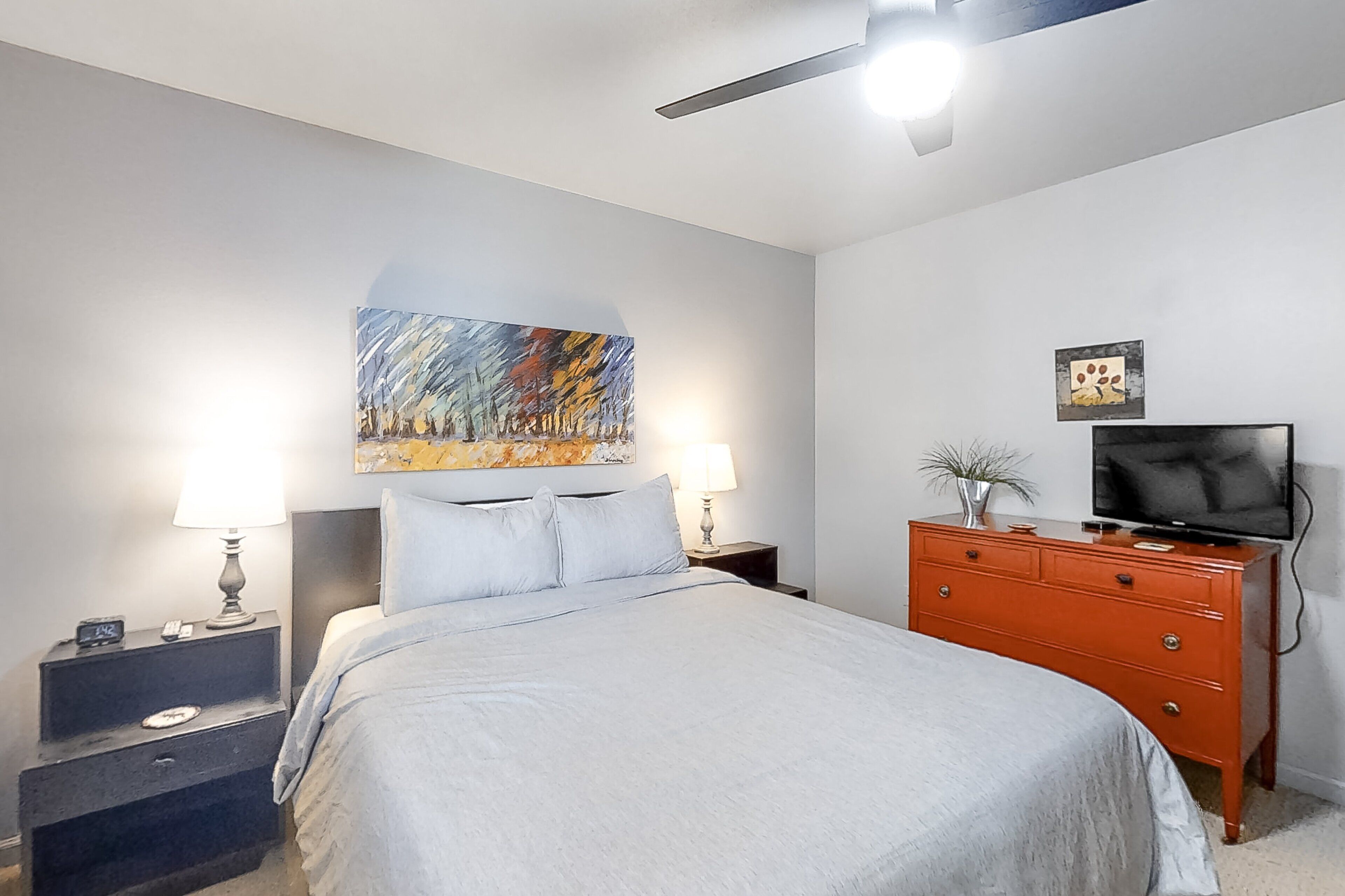 Un dormitorio con una cama, mesitas de noche, un aparador, un televisor y un cuadro en la pared.