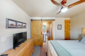 Ein Schlafzimmer mit einem Bett, einem Fernseher, einer Holzkommode, einem Deckenventilator und einem Badezimmer mit Waschbecken und Spiegel.