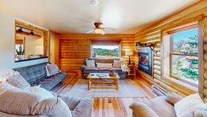 Interior de una cabaña de troncos con un sofá, sillones, una mesita de centro, chimenea y ventanas grandes que ofrecen vistas.