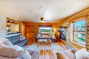 Ein Blockhaushaus-Interieur mit einer Couch, Sesseln, einem Couchtisch, einem Kamin und großen Fenstern mit Blick.