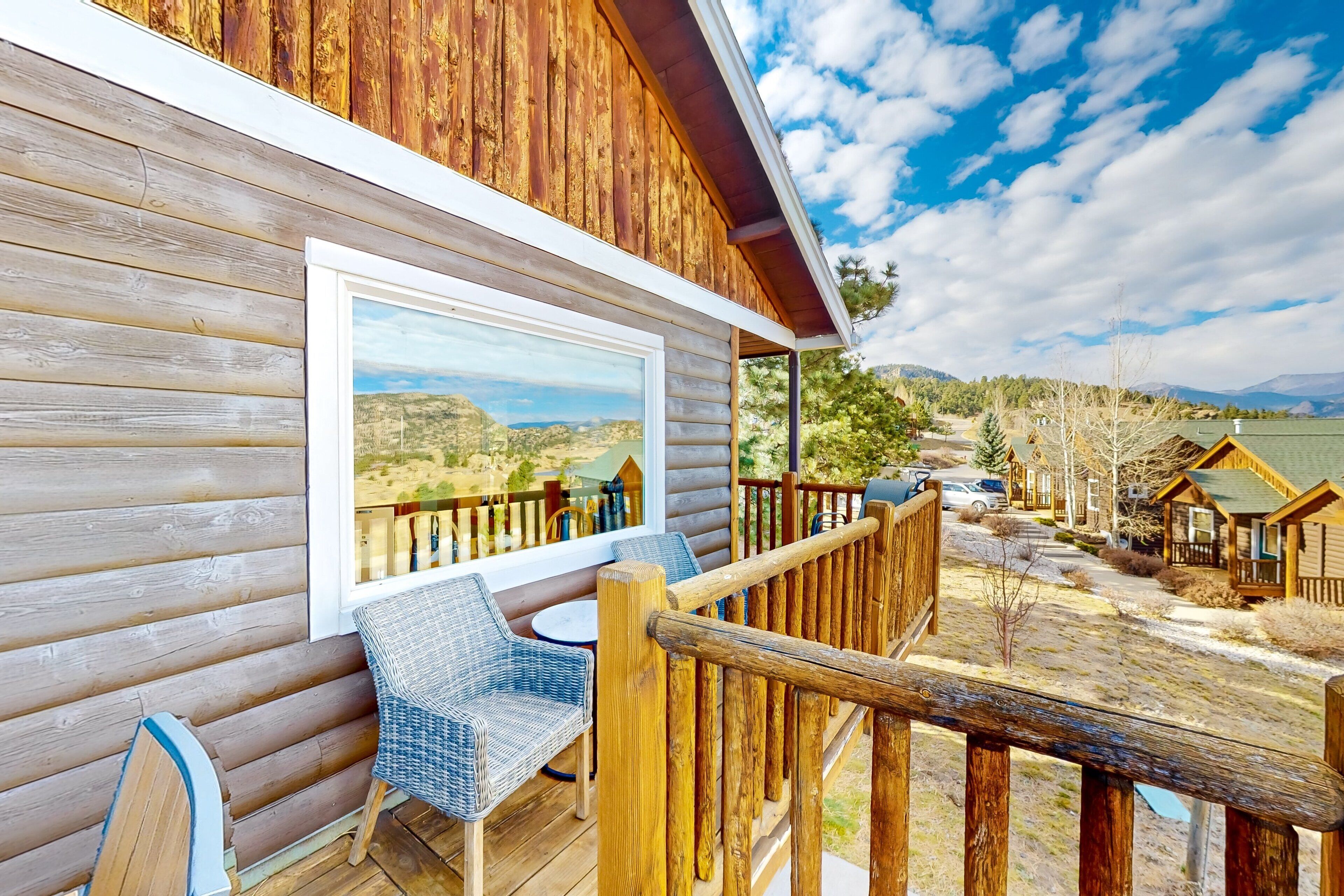 A wooden cabin with a deck, wicker chairs, and a large window overlooking a scenic landscape.