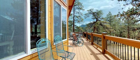 A wooden deck with metal chairs, large windows, and a view of trees and mountains.