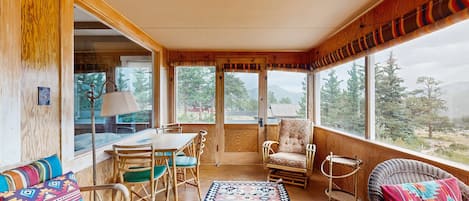 Un porche cubierto con muebles de madera, una alfombra estampada y amplias ventanas que dan a vistas de árboles y montañas.