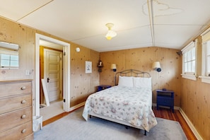 Ein Schlafzimmer mit Holzwand, einem Bett mit floralem Überwurf, einem Schminktisch, einem Nachttisch mit Lampe und einem Deckenventilator.