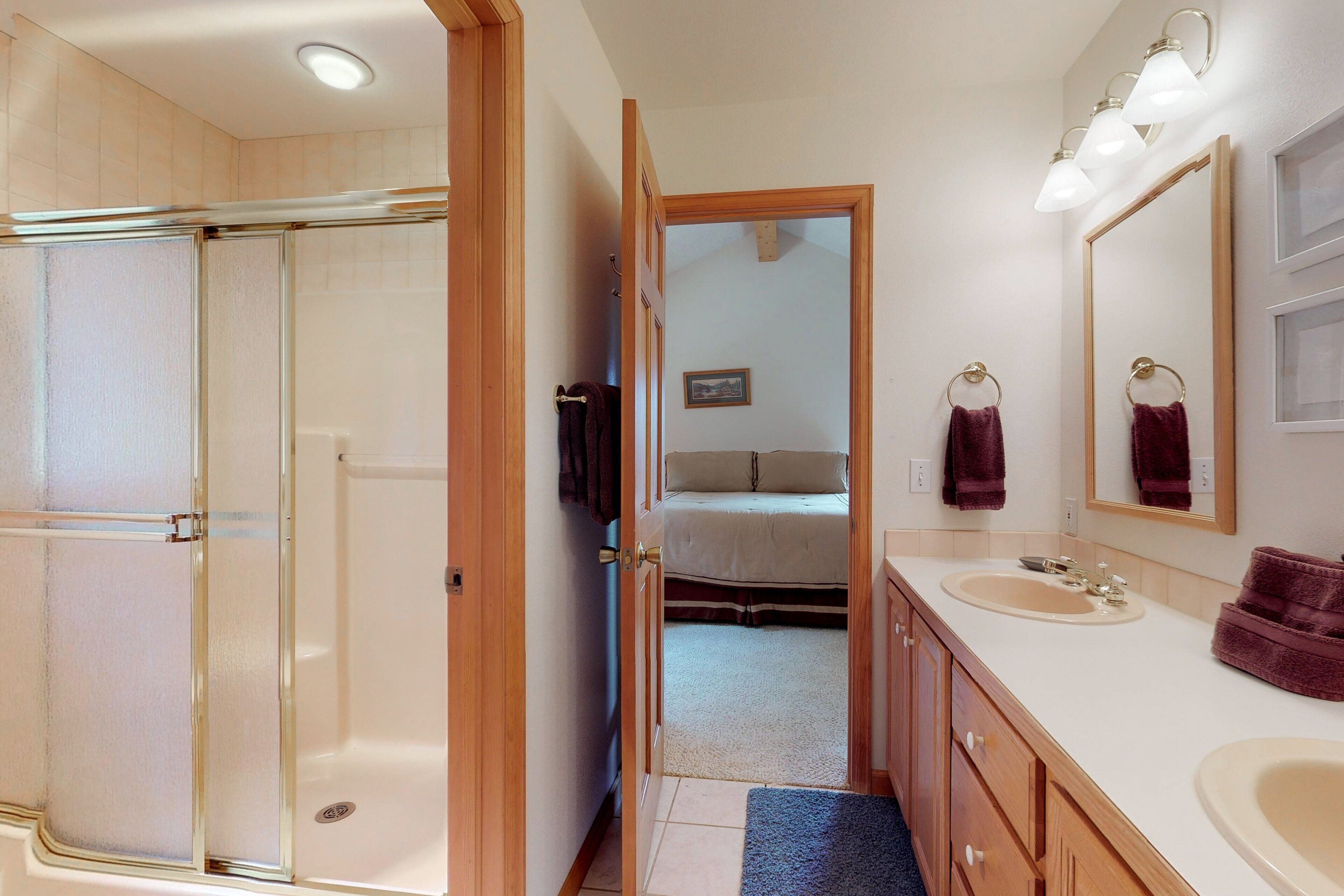 Een badkamer met een douche, twee wastafels, een spiegel en een deur naar een slaapkamer.