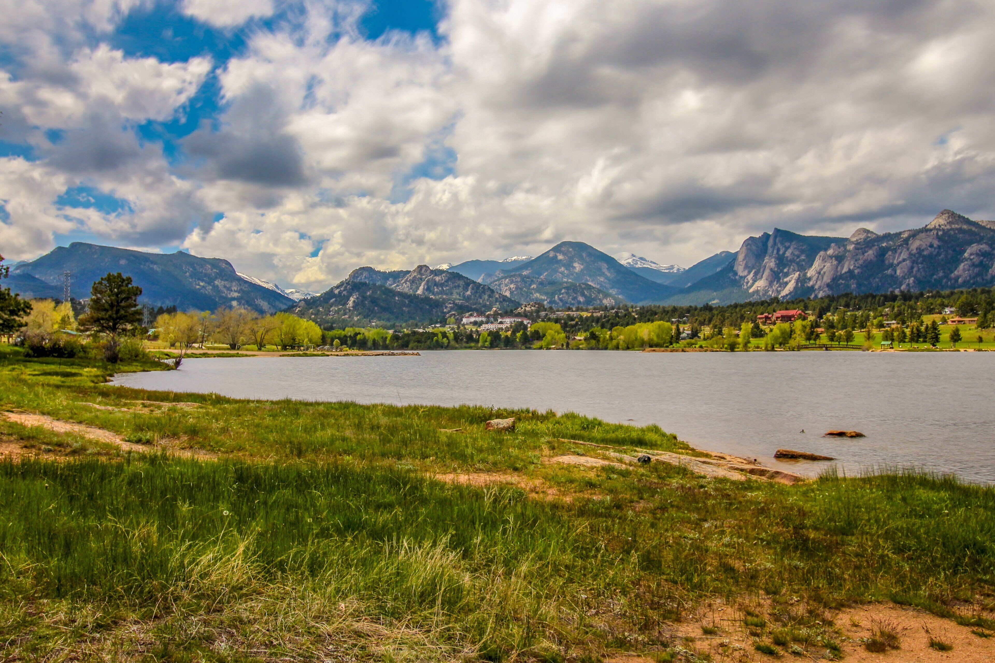 A lake with mountains in the background, surrounded by greenery and a few buildings.