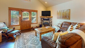 A spacious living room with a fireplace, a television mounted above it, a large window with blinds, and a ceiling fan. - Luxurious Dog-friendly Townhome Near Lake Estes With Fantastic Views (Estes Park)