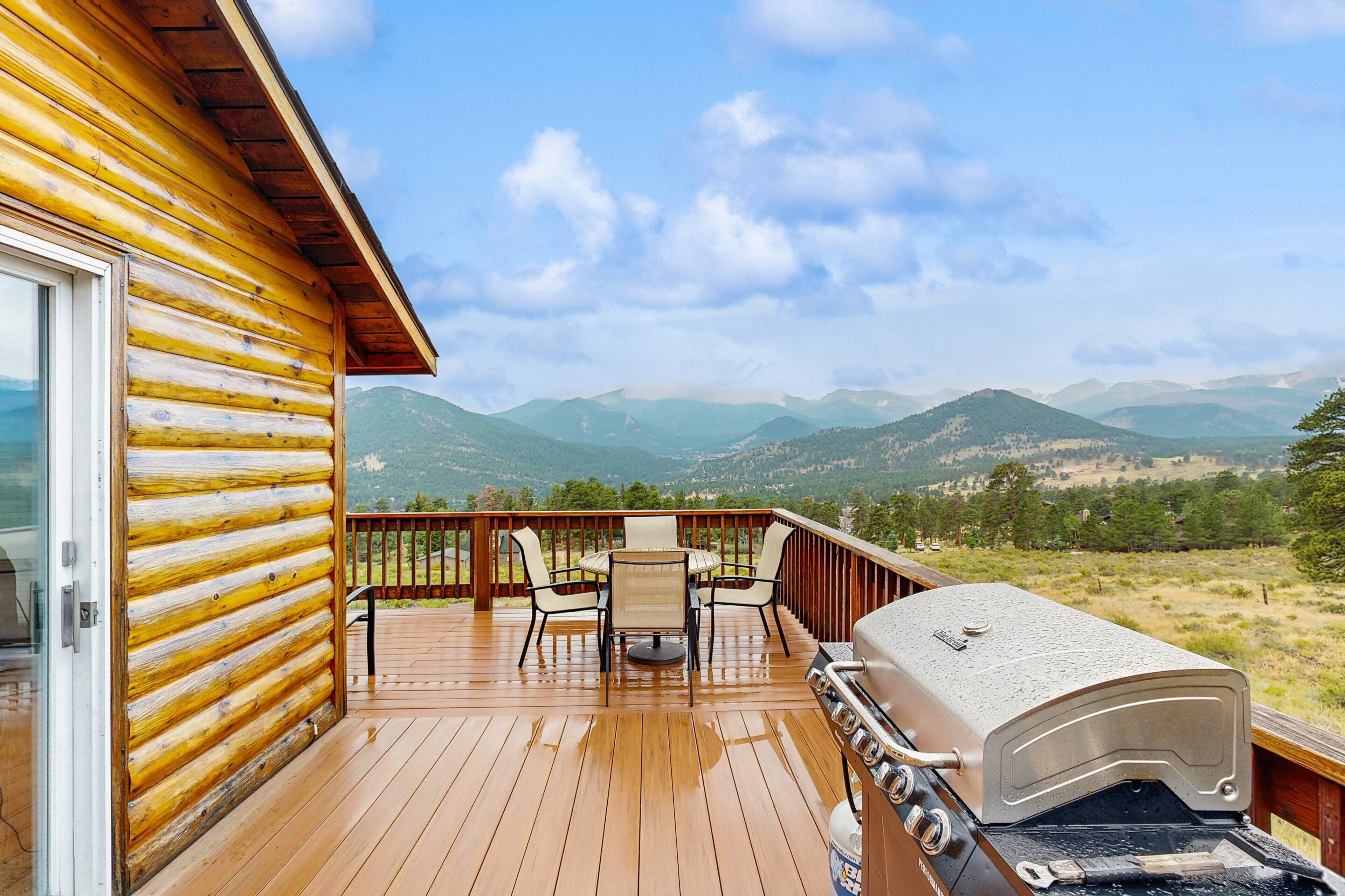 A wooden deck with a grill, chairs, and a table overlooking a mountainous landscape.