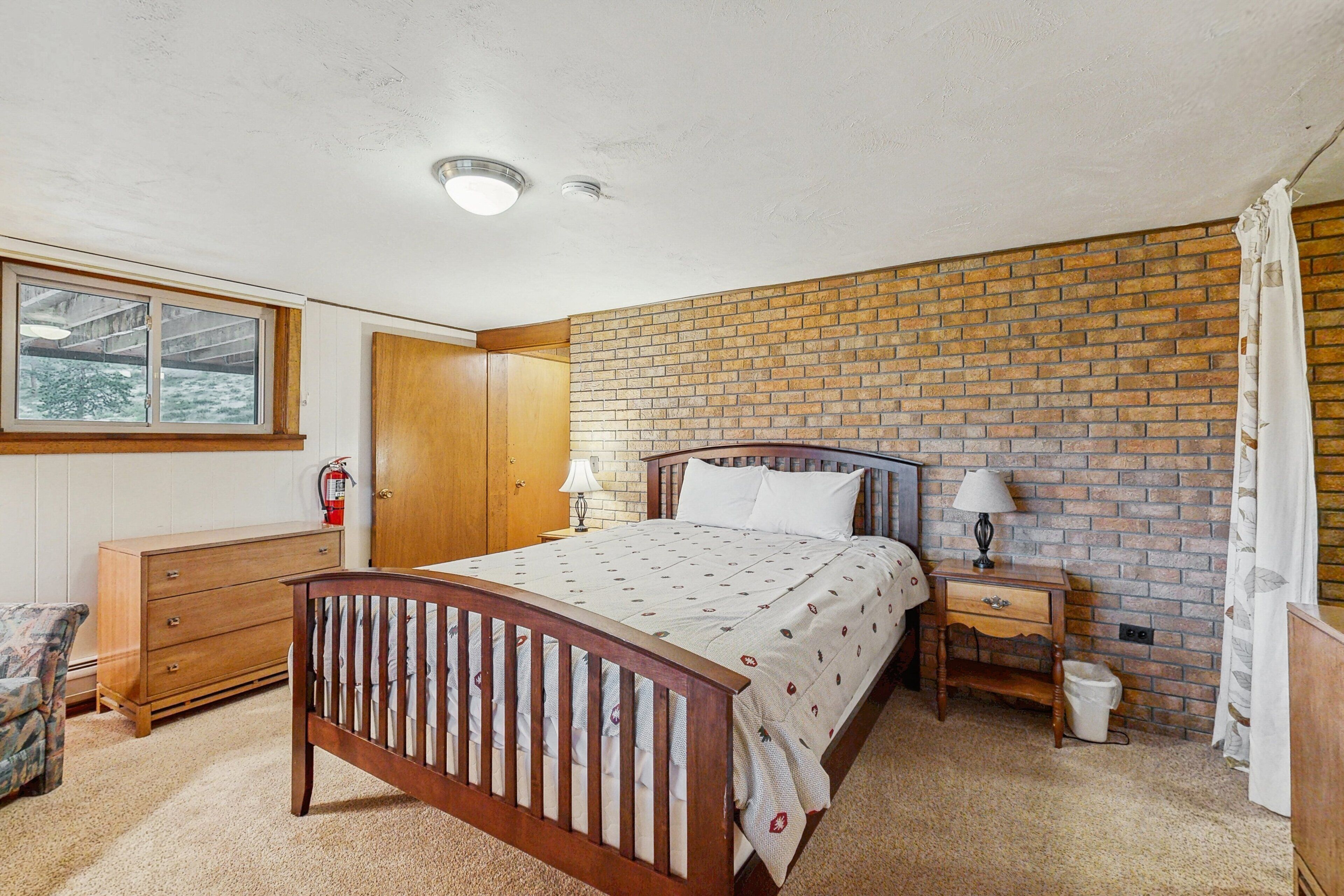 A bedroom with a wooden bed, a brick wall, a window, a wooden dresser, a nightstand, and a lamp.