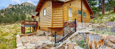 A log cabin with a stone-paved entrance, a wooden deck, and a mountain backdrop.