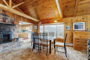 Ein rustikales Holzinterieur mit Kamin aus Stein, Esstisch mit Stühlen und Blick auf Berge durch große Fenster.