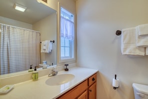 A bathroom with a white sink, wooden cabinets, a mirror, a window with a curtain, and a toilet with a towel rack. - Relaxing Condo in Estes Park w/ a Full Kitchen, gas Fireplace, & Furnished Deck (Estes Park)