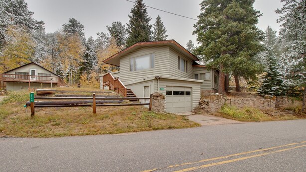 A two-story cabin with a garage, a wooden deck, and a stone wall. - Dog-friendly Mountain Haven With Full Kitchen, Furnished Patio, & Forest Views (Lyons)