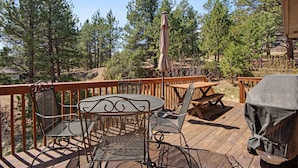 A wooden deck with outdoor furniture, including a table and chairs, surrounded by pine trees. - Dog-friendly, Mountain-view Home With Deck & Grill (Estes Park)