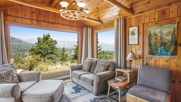 Ein gemütliches Zimmer mit Holzbalkendecke, großen Fenstern mit Bergblick, einer Couch, einem Sessel und einem kleinen Tisch mit Lampe.