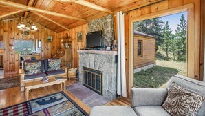 Ein Blockhaus-Interieur mit Kamin aus Stein, Holzmöbeln und Blick auf den umliegenden Wald.
