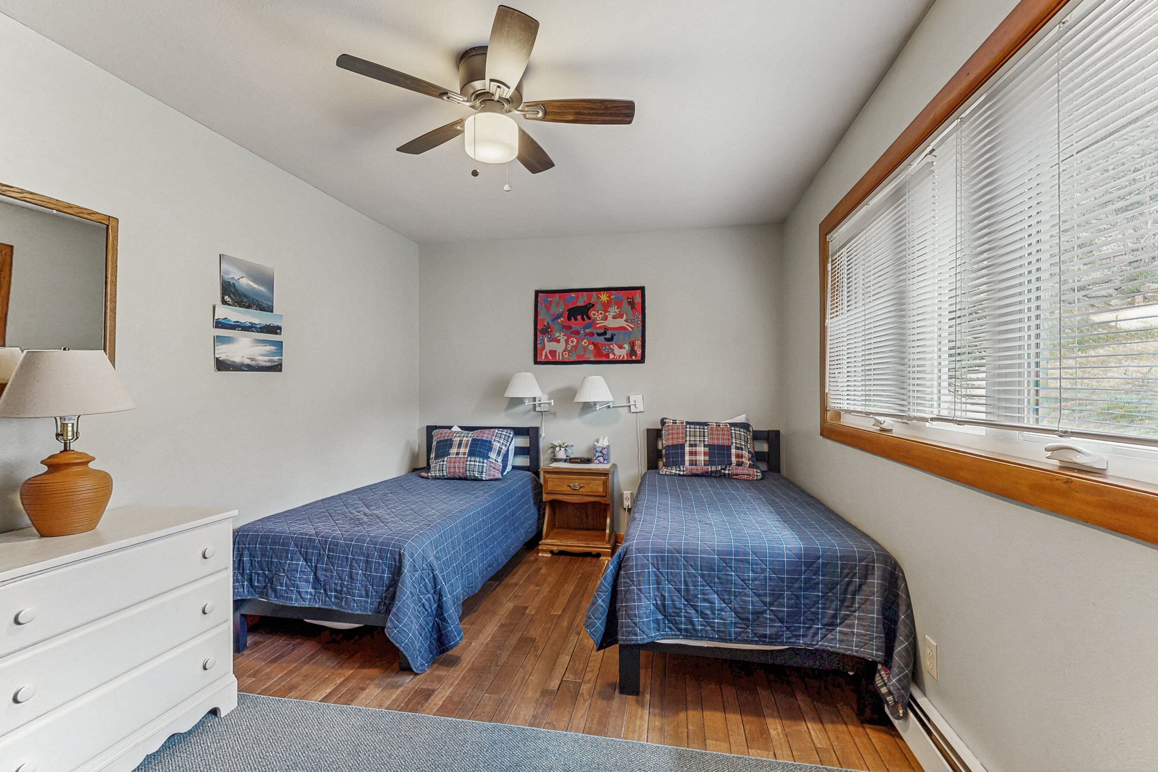 A bedroom with two beds, a ceiling fan, a dresser, a window with blinds, and artwork on the walls.