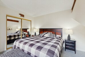 A bedroom with a large mirror, a bed with a checkered bedspread, two bedside tables with lamps, and a small pine tree decoration. - Charming Condo With Sunroom & Patio - Near Golf, Town & National Park (Estes Park)