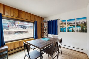 A dining area with a wooden table and chairs, a view through a window, and a shelf with decorative items. - Cozy Cottage With Grill Near River and Downtown (Estes Park)