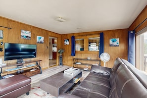 A living room with a leather sofa, a flat-screen TV, wooden walls, and artwork. - Cozy Cottage With Grill Near River and Downtown (Estes Park)