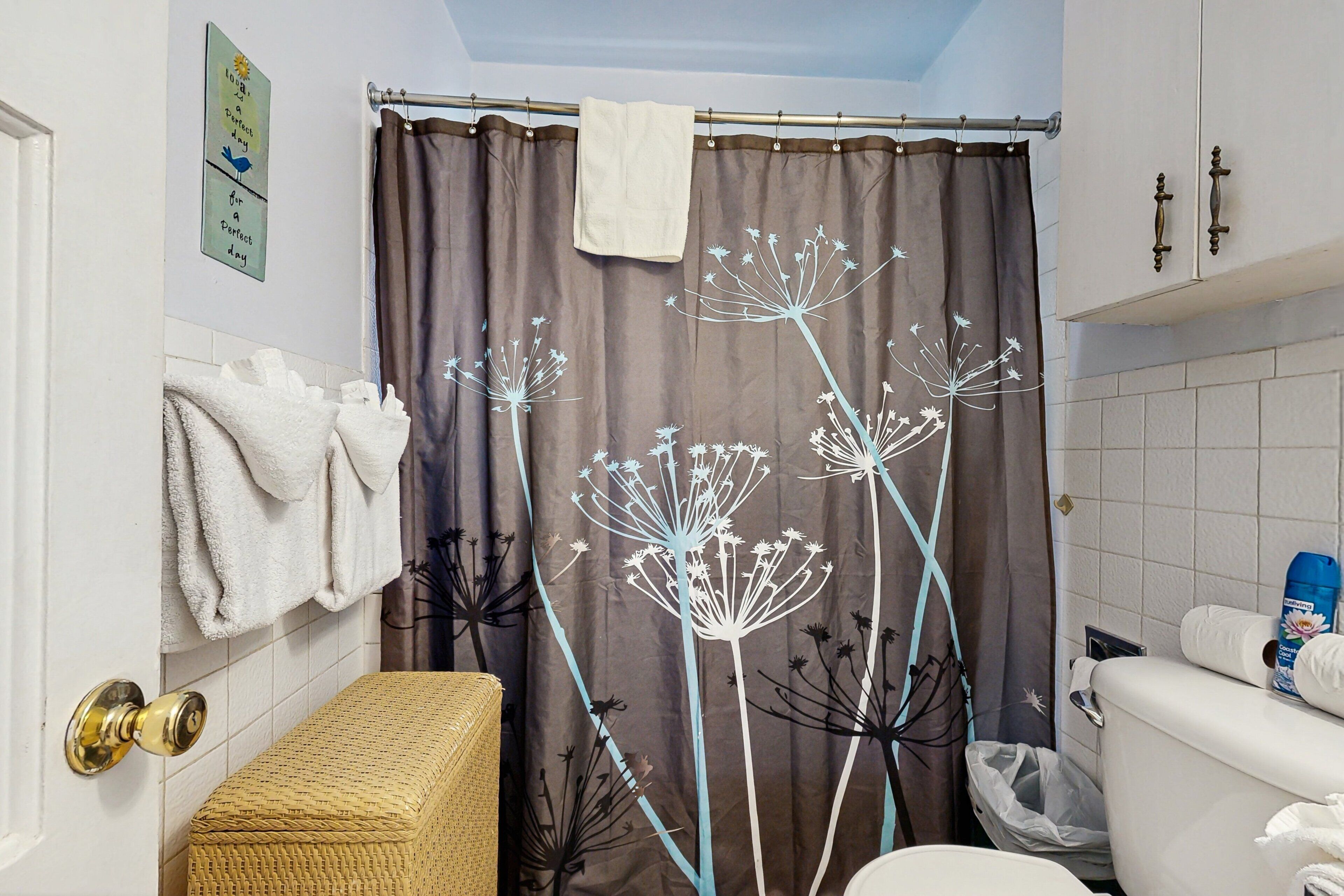 A bathroom with a shower curtain featuring a dandelion design, a white toilet, a trash bin, and a wicker bench.