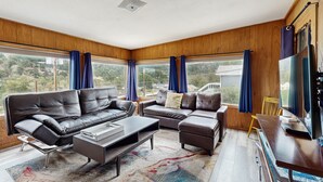 Una sala de estar moderna con un sofá modular, una mesa de centro y un televisor sobre una mesa de madera. Amplias ventanas con cortinas ofrecen vistas al exterior.