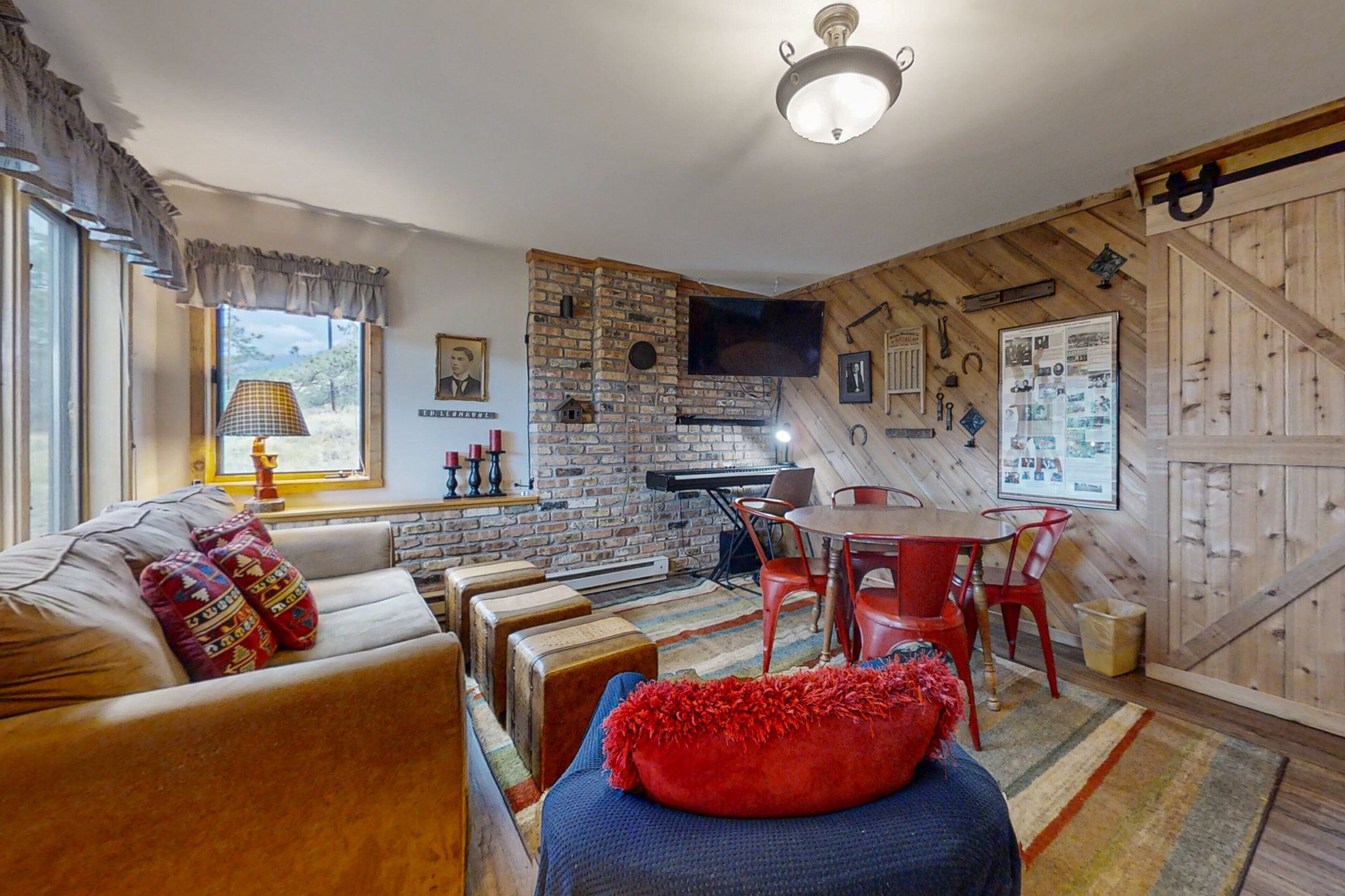 A cozy living room with a brick wall, a flat-screen TV, a piano, and a dining area with red chairs.