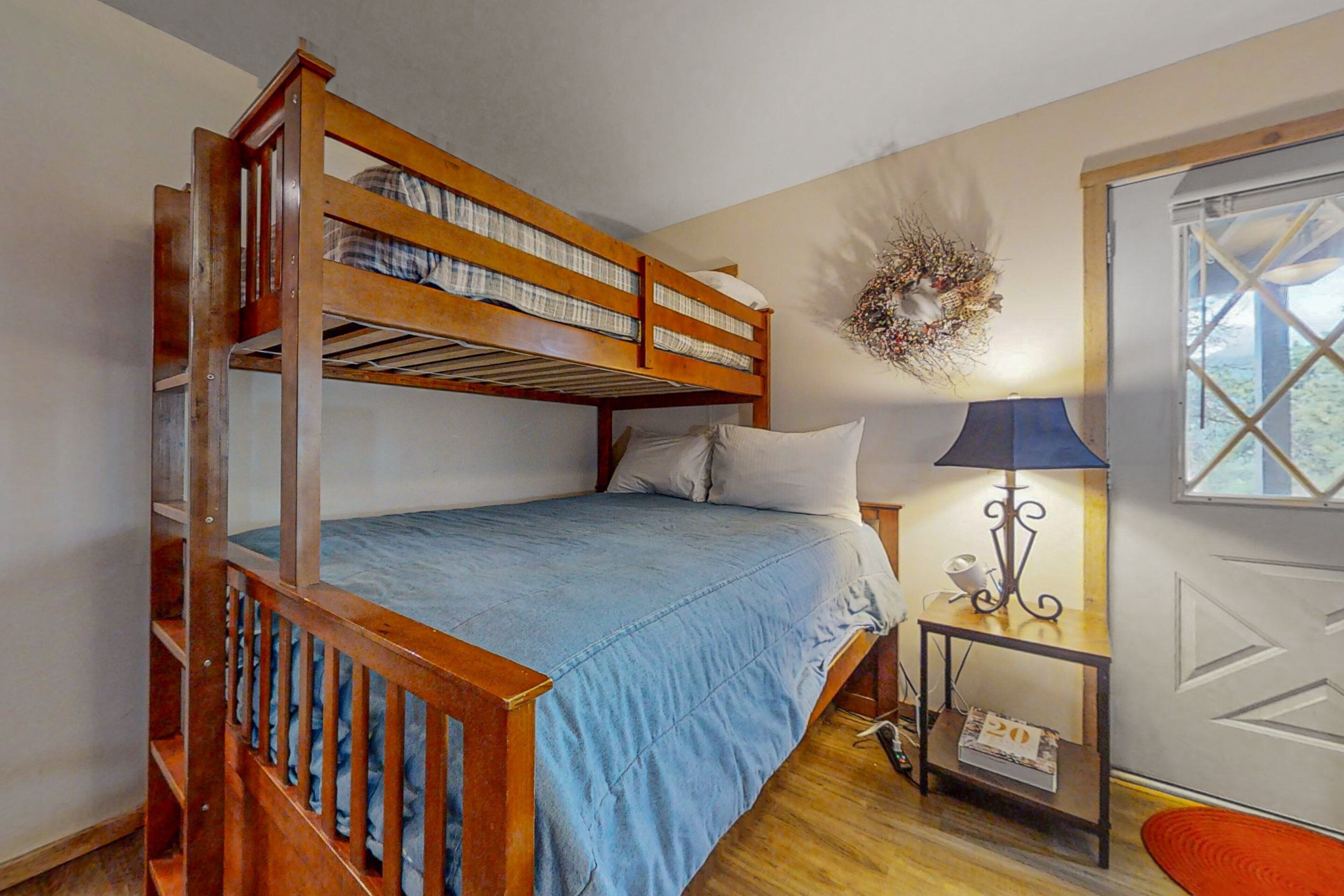 A bunk bed room with a blue bedspread, a small table with a lamp, and a decorative wreath on the wall.