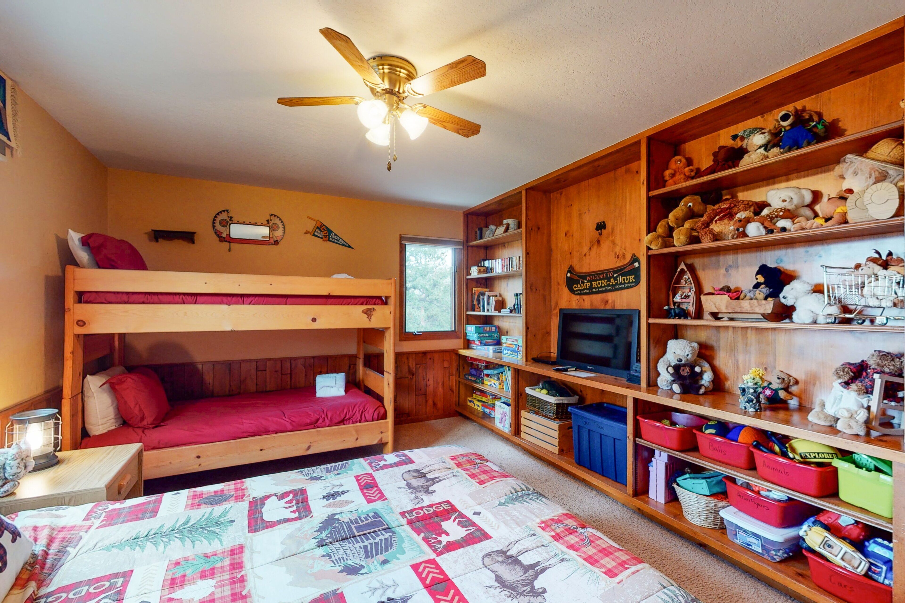 A bunk bed room with a ceiling fan, wooden shelves with toys, and a bookshelf.