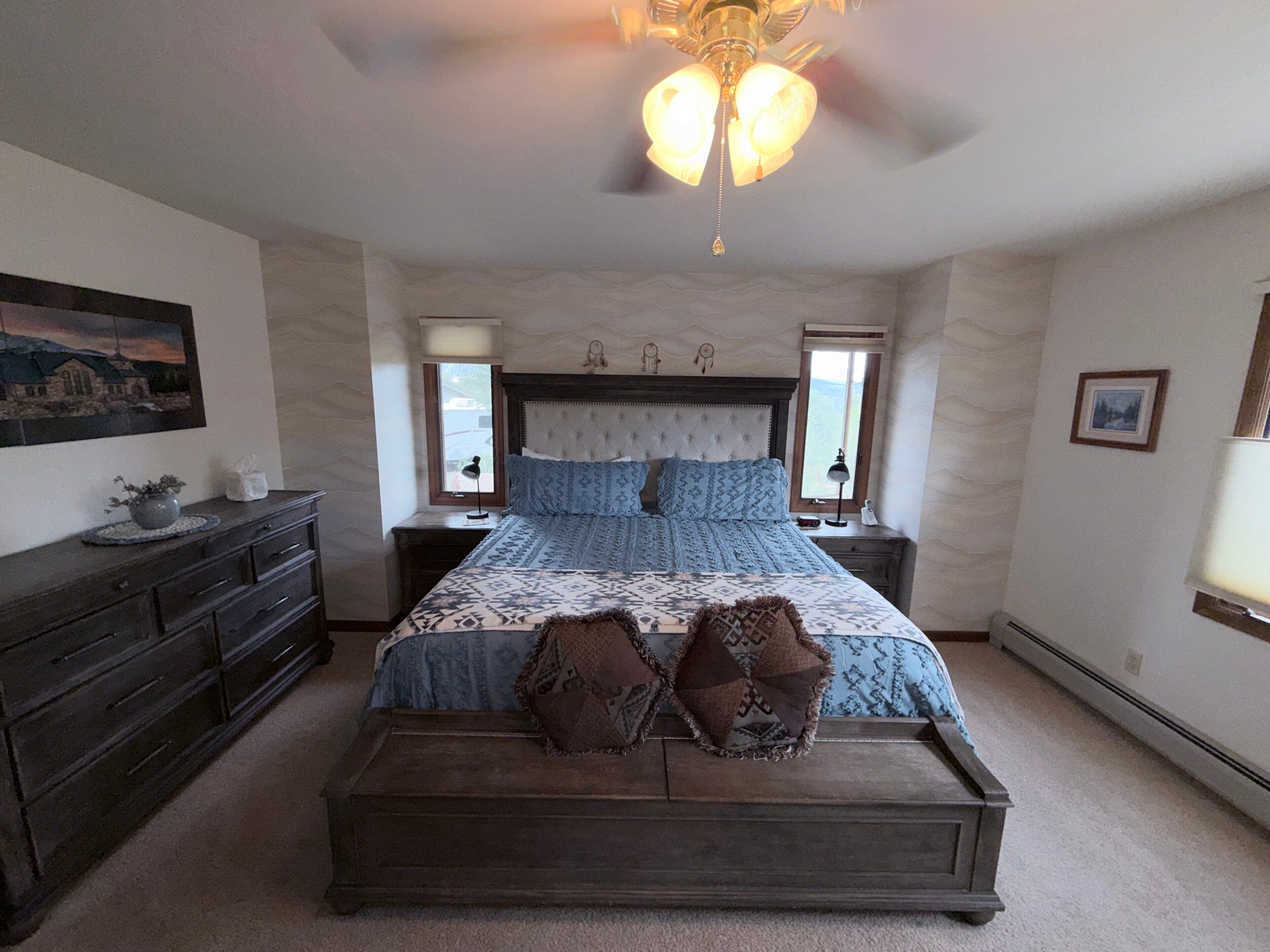 A bedroom with a large bed, a dresser, a nightstand, a lamp, and a ceiling fan.