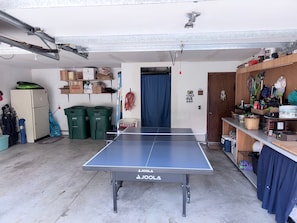 Ein Pingpongtisch in einer Garage mit Kühlschrank, Aufbewahrungsboxen und diversen Gegenständen auf Regalen.