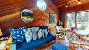 Ein gemütliches Blockhaus-Interieur mit Holzbalkendecke, einem blauen Sofa mit gemusterten Kissen, einem Esstisch und Stühlen.