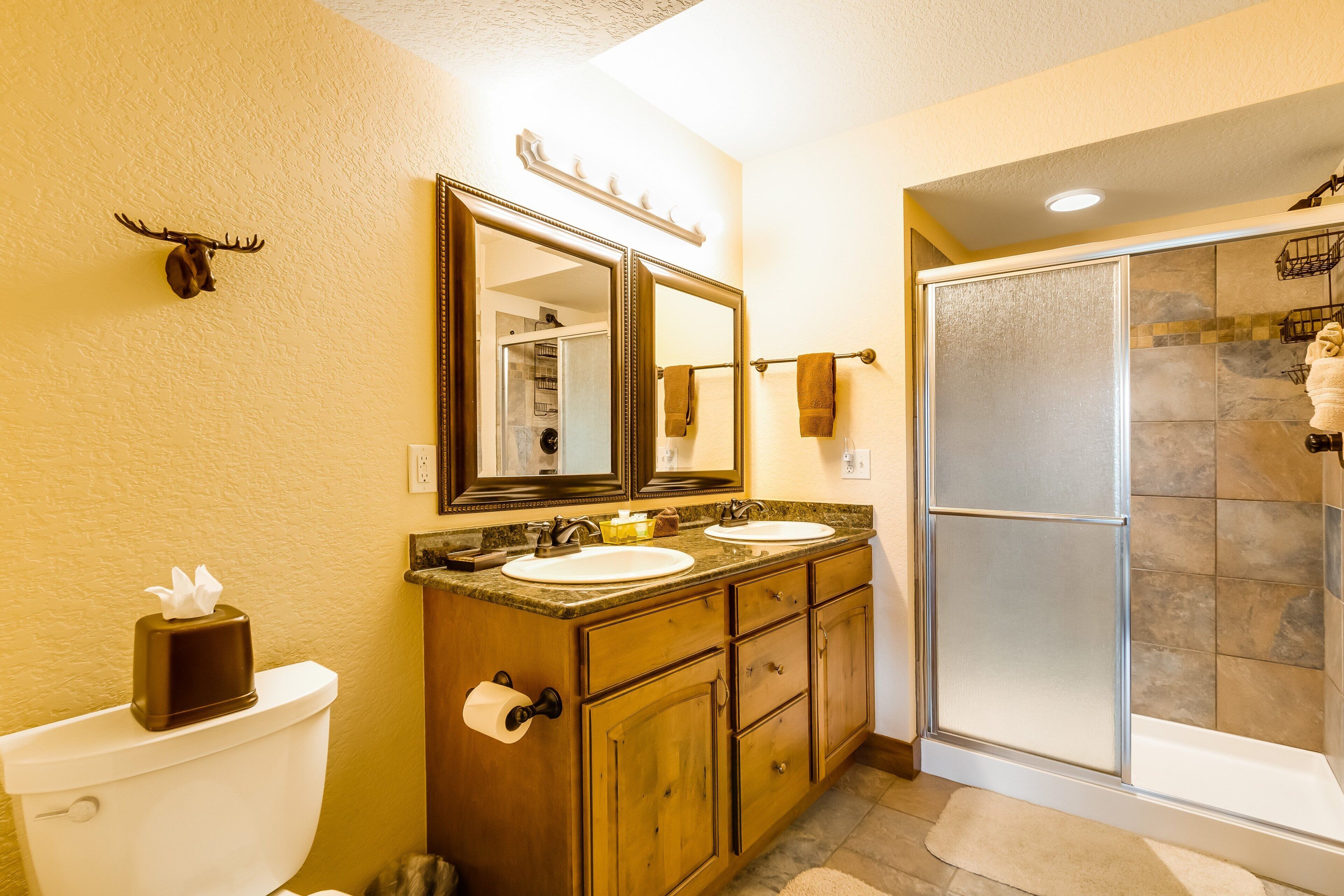 A bathroom with a toilet, double sinks, a mirror, a shower enclosure, and a mounted deer head.