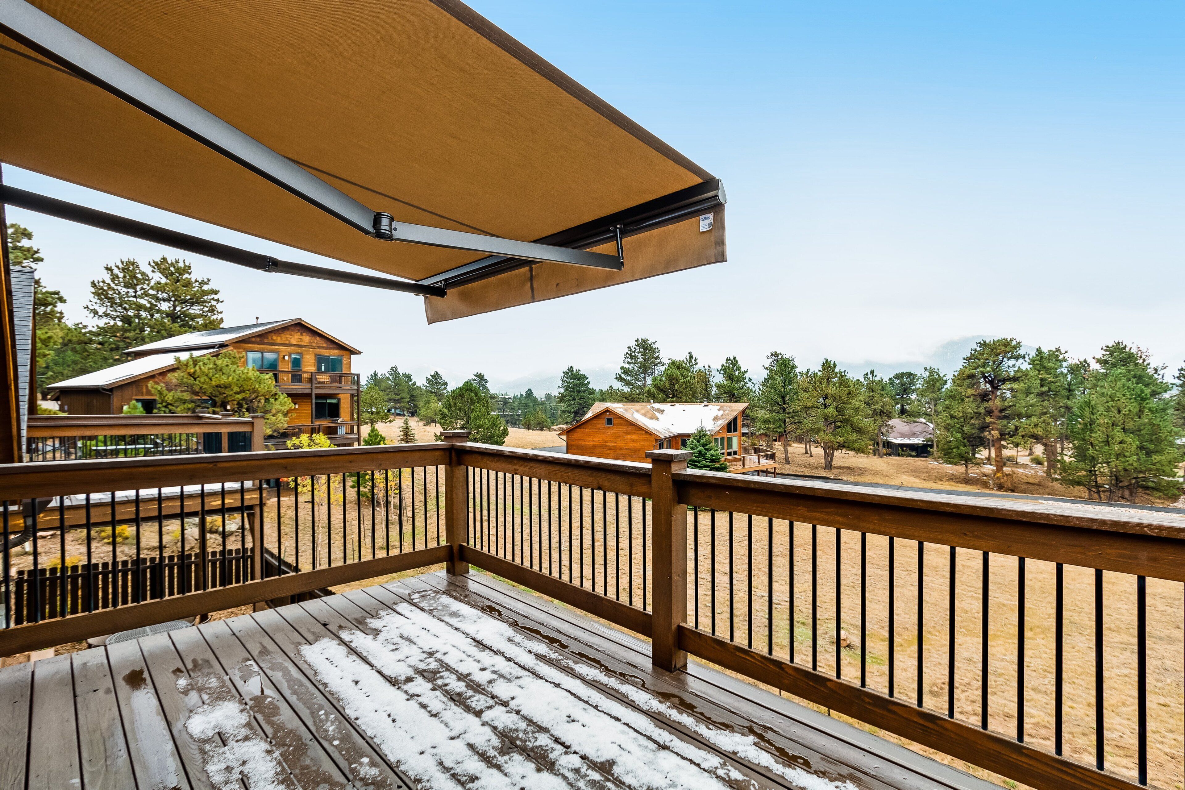 Um deck de madeira com guarda-corpo de metal, um toldo retrátil e vista para uma área residencial com casas e árvores.
