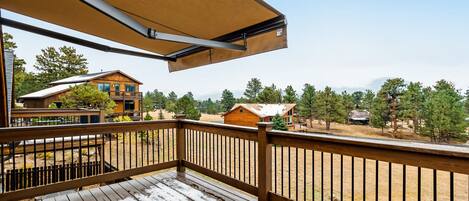 Um deck de madeira com guarda-corpo de metal, um toldo retrátil e vista para uma área residencial com casas e árvores.