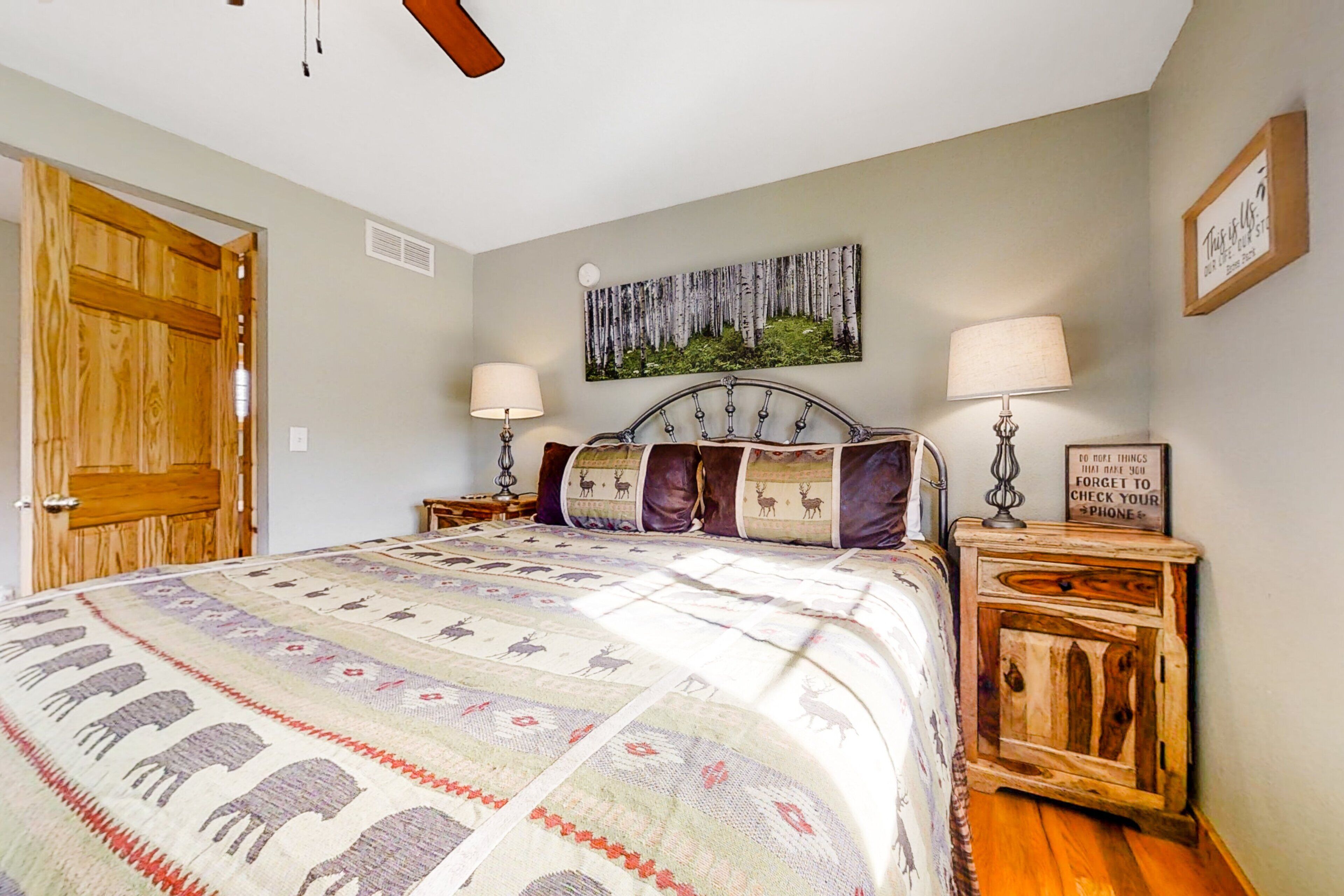 A bedroom with a bed, two bedside tables, a wooden door, a wall-mounted picture, and a ceiling fan.