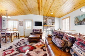 Ein großzügiges Wohnzimmer mit Holzboden, einem Steinkamin, einem großen Sofa mit gemusterten Kissen und einem Essbereich mit Holzmöbeln.