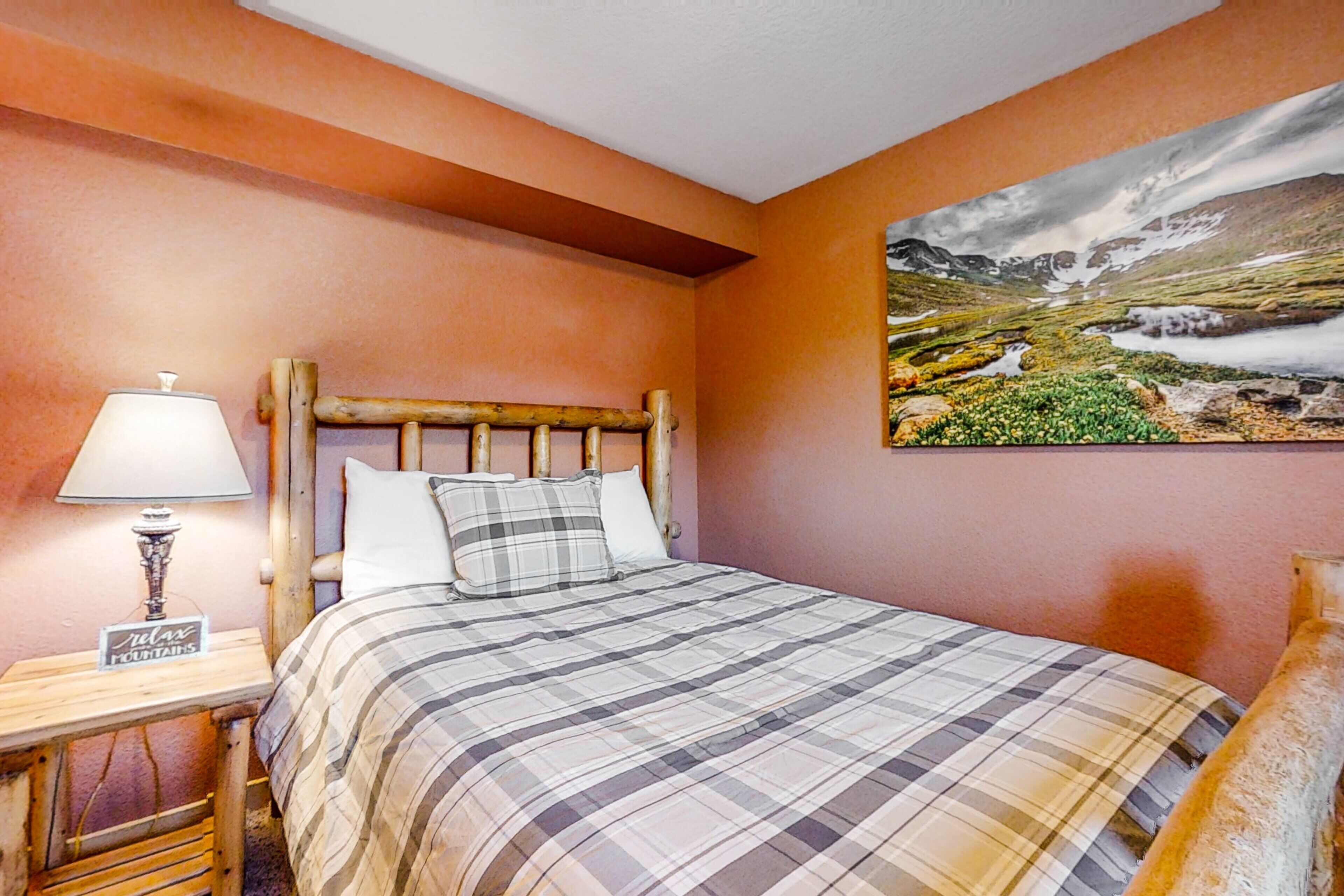 A bedroom with a wooden headboard, a plaid bedspread, a nightstand with a lamp, and a painting of a landscape on the wall.