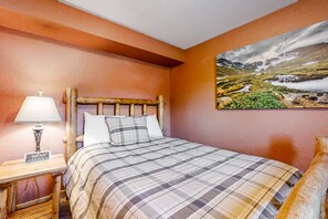 Ein Schlafzimmer mit einem Holz-Kopfteil, einer karierten Bettdecke, einem Nachttisch mit Lampe und einem Landschaftsbild an der Wand.
