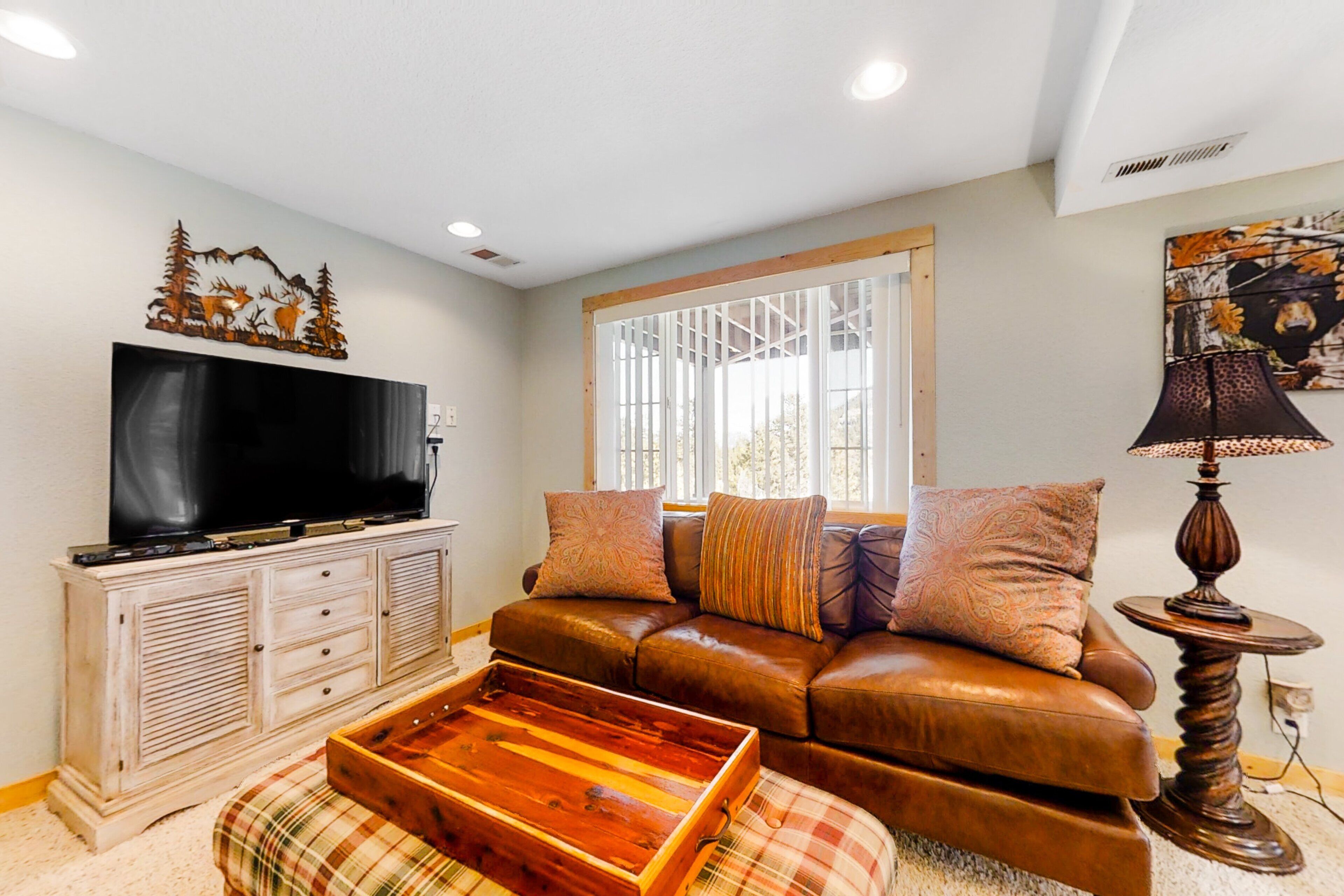 A living room with a television, a brown leather sofa, a plaid ottoman, a wooden cabinet, and a window with blinds.