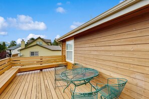 Um deck de madeira com uma mesa e cadeiras de metal, um cercado de madeira e uma casa ao fundo.