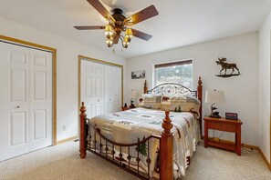 Quarto com cama, ventoinha no teto, mesa de cabeceira, luminária, janela e um adorno na parede.