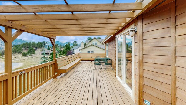 Una terraza de madera con un porche, una mesa con sillas y vistas a casas y montañas.
