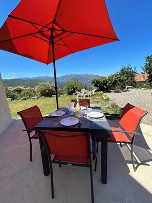 An outdoor dining area with a table set for two, a red umbrella, and a scenic mountain view in the background.