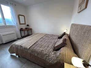 A bedroom with a bed, bedside lamp, window, radiator, and a small table with a lamp.
